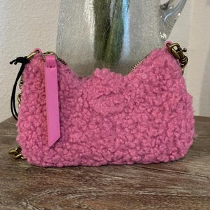 May include: A small, fluffy pink handbag with a gold chain strap and zipper closure. The bag has a textured, curly exterior and a pink leather zipper pull. The bag is sitting on a wooden surface.