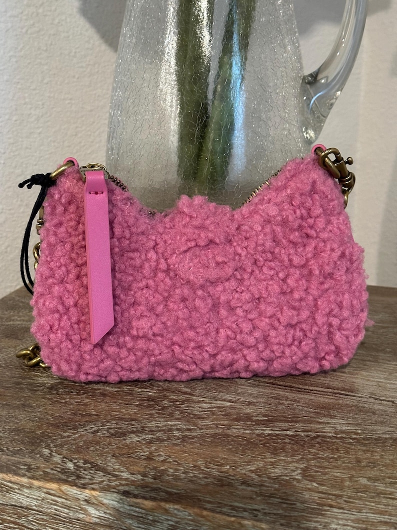 May include: A small, fluffy pink handbag with a gold chain strap and zipper closure. The bag has a textured, curly exterior and a pink leather zipper pull. The bag is sitting on a wooden surface.