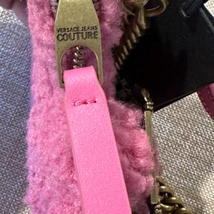 May include: Close-up of a pink Versace Jeans Couture handbag. The bag features a gold-toned zipper with the brand's name, a pink leather strap, and a gold chain strap. The bag's material has a fuzzy texture, and a black tag is attached.