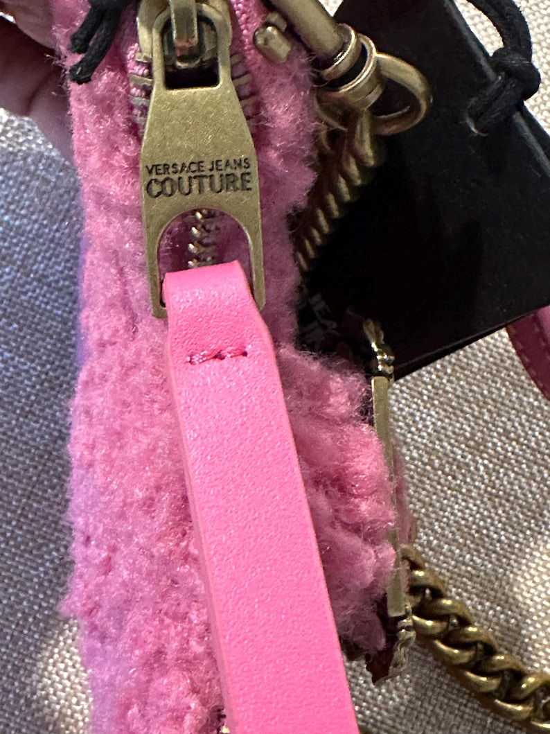 May include: Close-up of a pink Versace Jeans Couture handbag. The bag features a gold-toned zipper with the brand's name, a pink leather strap, and a gold chain strap. The bag's material has a fuzzy texture, and a black tag is attached.