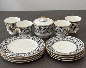 May include: A tea set featuring four white porcelain cups with black floral patterns and gold trim, a matching lidded sugar bowl, and eight saucers. The cups have gold-toned handles, and the saucers and sugar bowl feature intricate black scrollwork.