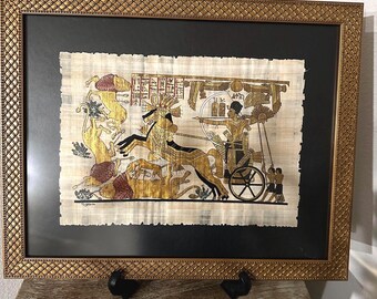 May include: Framed papyrus art depicting an Egyptian pharaoh riding a chariot pulled by horses. The artwork is done in gold and black on a beige background.