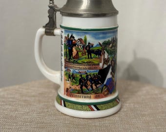 May include: A white ceramic beer stein with a pewter lid and a handle. The stein features a colorful scene of people in traditional clothing and a landscape. The text "Erinnerung an" is printed on the stein.