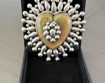 May include: A silver-toned brooch in a black box. The brooch features a heart-shaped center with a pearlescent finish, surrounded by a silver design. The brooch is displayed in an open black jewelry box.