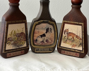 May include: Three vintage Beam's Choice whiskey bottles. Each bottle features a different animal scene: a raccoon, a dog, and a fox. The bottles are brown with a dark brown cap and a rectangular label with gold trim.