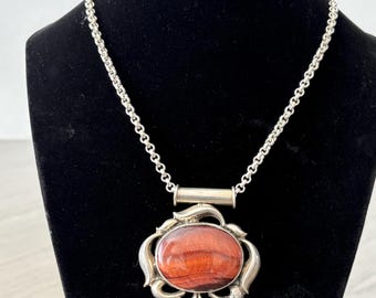 May include: Silver-toned necklace with a large, oval pendant. The pendant features a polished, reddish-brown stone set within an ornate, floral-inspired frame. The chain is a delicate link style, and the necklace is displayed on a black mannequin.
