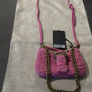 May include: A pink, fluffy shoulder bag with a gold chain strap and a matching pink leather strap. The bag has a gold clasp and a tag that reads "VERSACE JEANS COUTURE".