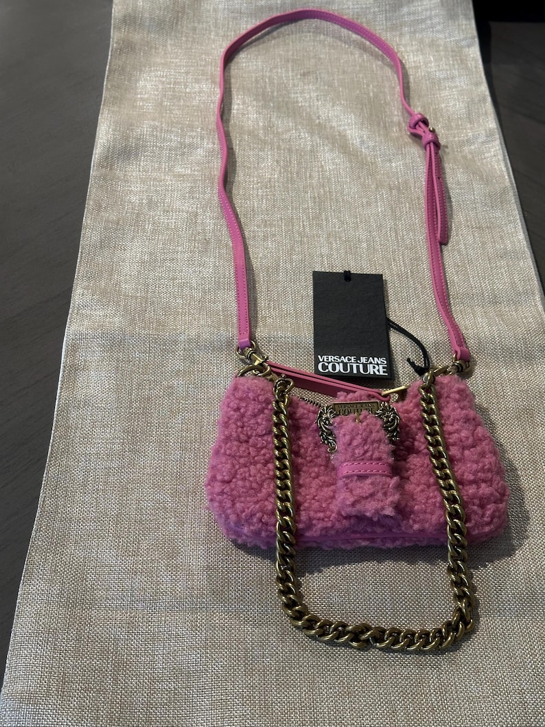May include: A pink, fluffy shoulder bag with a gold chain strap and a matching pink leather strap. The bag has a gold clasp and a tag that reads "VERSACE JEANS COUTURE".