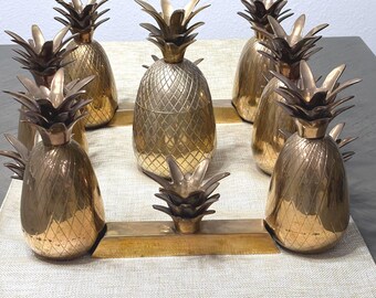 May include: A collection of gold-colored pineapple-shaped objects. The pineapples have textured surfaces and detailed leafy crowns. They are arranged on a rectangular gold-colored base, creating a decorative centerpiece.