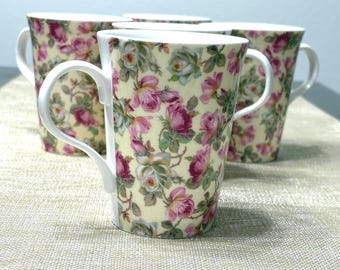 May include: Four white ceramic mugs with a floral pattern of pink roses and green leaves on a cream background. Each mug has a curved handle. The mugs are arranged on a light-colored surface.