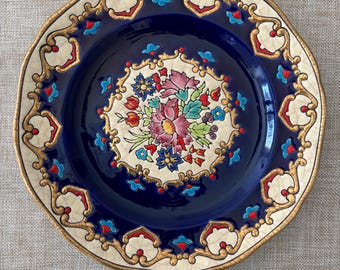 May include: A decorative plate with a dark blue center and a floral design. The plate features a cream-colored border with gold accents and a repeating pattern. The floral design in the center includes pink, red, and blue flowers.
