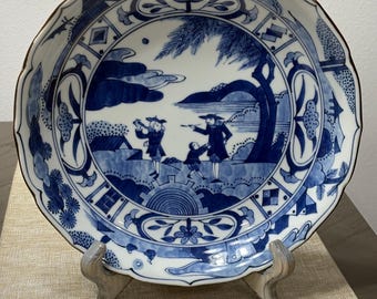 May include: A blue and white ceramic plate with a scalloped edge and a brown rim. The plate features a detailed landscape scene with figures and trees. Decorative floral and geometric patterns adorn the border. The plate is displayed on a wooden stand.