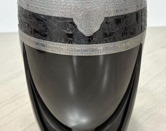 May include: A black glass vase featuring a wide silver-toned metal band around the upper rim. The metal band is decorated with intricate embossed floral and geometric patterns. The vase has a smooth, rounded body supported by three curved legs.