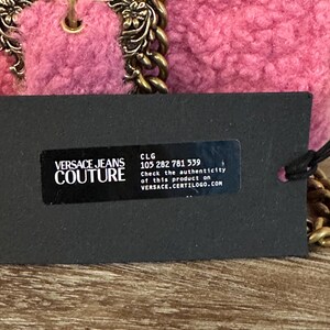 May include: A pink Versace Jeans Couture handbag with a gold chain strap and hardware. The bag has a textured, plush exterior. A black tag displays the brand's logo and product authentication information.