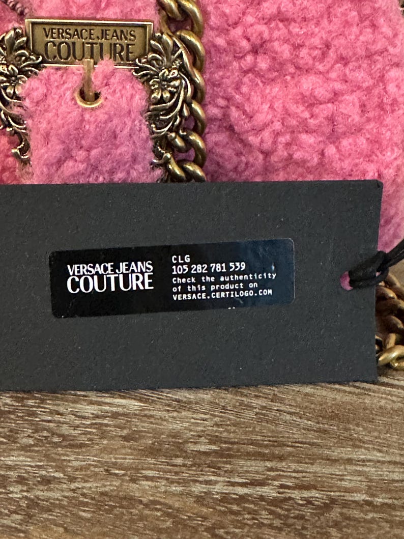 May include: A pink Versace Jeans Couture handbag with a gold chain strap and hardware. The bag has a textured, plush exterior. A black tag displays the brand's logo and product authentication information.
