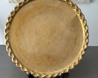 May include: A gold-colored, round decorative tray with an ornate floral design in the center. The tray has a scalloped edge and is displayed on a black stand. The tray is likely made of metal and is suitable for home decor.