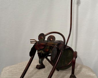 May include: A whimsical metal cat sculpture with a long, curled tail. The cat is made of brown and green metal and has a playful expression. The sculpture is standing on a light brown fabric surface.