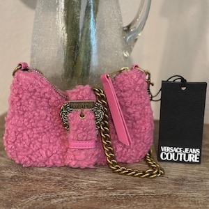May include: A pink, textured shoulder bag with a gold chain strap and a decorative buckle. The bag has a matching pink handle and a black tag with the "VERSACE JEANS COUTURE" logo.
