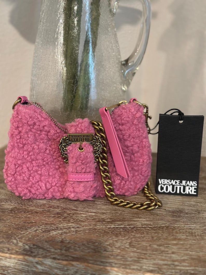May include: A pink, textured shoulder bag with a gold chain strap and a decorative buckle. The bag has a matching pink handle and a black tag with the "VERSACE JEANS COUTURE" logo.