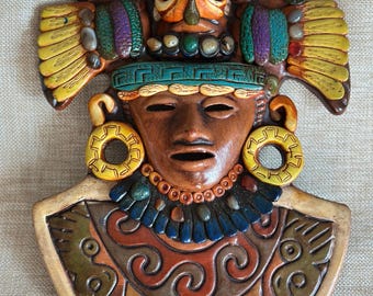 May include: A colorful, handcrafted wall mask depicting an ancient figure. The mask features a brown face with closed eyes, yellow earrings, and a detailed headdress with various colors, including yellow, green, purple, and blue. The mask has a detailed collar and a beige border.