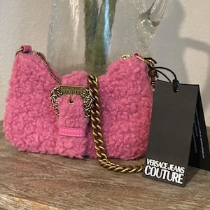 May include: A pink, fluffy Versace Jeans Couture shoulder bag with a gold chain strap and buckle. The bag has a rectangular shape and a small leather strap. A black tag with the brand's logo hangs from the bag.