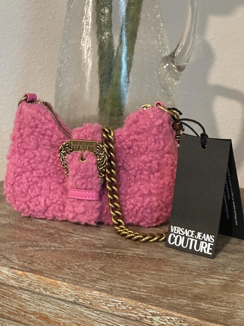 May include: A pink, fluffy Versace Jeans Couture shoulder bag with a gold chain strap and buckle. The bag has a rectangular shape and a small leather strap. A black tag with the brand's logo hangs from the bag.