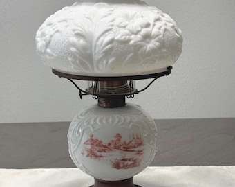 May include: A white milk glass oil lamp featuring a large, embossed floral shade and a smaller, spherical base decorated with a reddish-brown landscape scene. The lamp has a clear glass chimney and a dark, ornate metal base with four feet.
