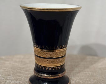 May include: A black and gold ceramic vase with a flared opening and a wide base. The vase has a gold trim around the top and two bands of gold floral designs around the middle.