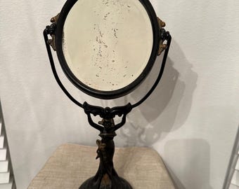 May include: An antique black vanity mirror with a round, aged mirror surface. The mirror is supported by a decorative stand featuring a sculpted female figure. The stand has a round base and ornate detailing with gold accents.