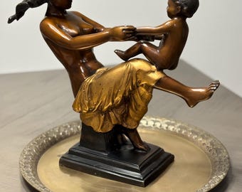 May include: A bronze sculpture depicting a woman holding a child. The woman is seated, with the child balanced on her lap. The sculpture has a gold-colored dress and is set on a decorative metal tray.