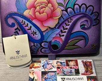 May include: Purple leather clutch with a floral design featuring a pink rose, blue paisley, and green leaves. The clutch has a small tag that reads "Anuschka Since 1988".