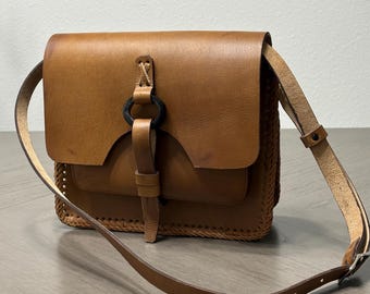 May include: A tan leather shoulder bag featuring a rectangular flap, a central leather strap secured by a black circular ring, and intricate braided leather detailing along the side panels. The bag includes an adjustable leather shoulder strap.