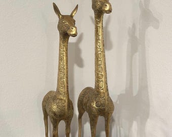 May include: Two gold-colored brass giraffe sculptures. The giraffes are standing on a beige surface. The larger giraffe is on the right and the smaller giraffe is on the left.