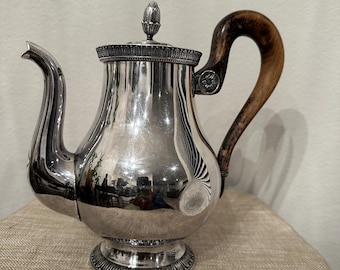 May include: A silver teapot with a wooden handle. The teapot has a round body and a decorative lid. The teapot is sitting on a beige surface.