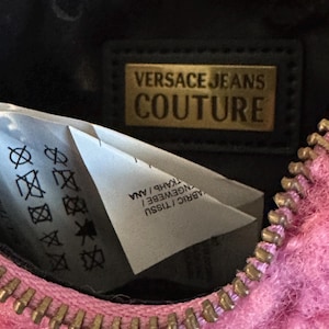May include: Close-up of a pink, fuzzy Versace Jeans Couture bag with a gold zipper. The interior is black, with a gold rectangular label that reads "VERSACE JEANS COUTURE". A white tag with care symbols is visible.