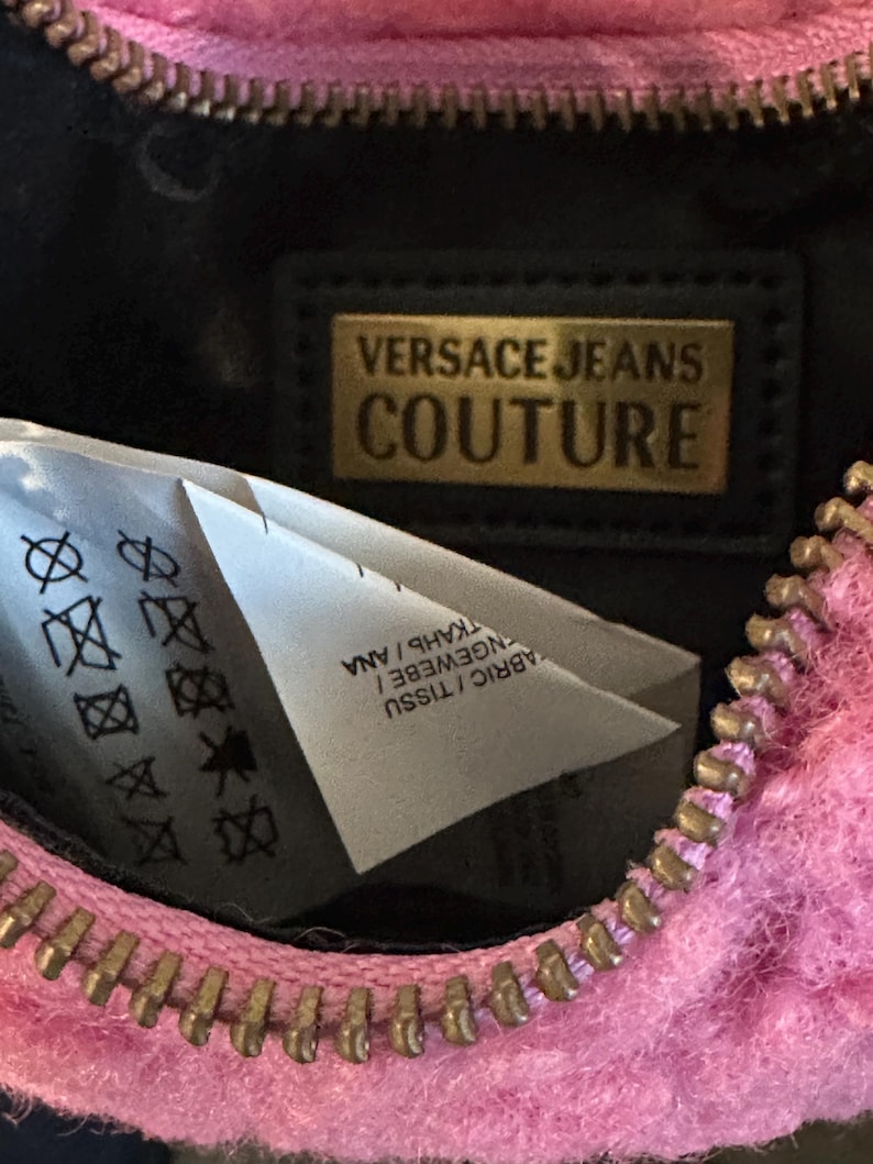May include: Close-up of a pink, fuzzy Versace Jeans Couture bag with a gold zipper. The interior is black, with a gold rectangular label that reads "VERSACE JEANS COUTURE". A white tag with care symbols is visible.