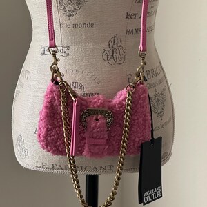 May include: A pink, fluffy crossbody bag with a gold chain strap and a pink leather strap. The bag has a gold buckle closure and a black tag that reads "VERSACE JEANS COUTURE."