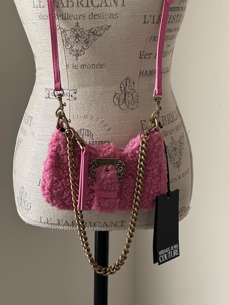 May include: A pink, fluffy crossbody bag with a gold chain strap and a pink leather strap. The bag has a gold buckle closure and a black tag that reads "VERSACE JEANS COUTURE."