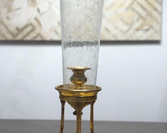 May include: A tall, clear glass vase with a textured surface and a gold rim, set on a decorative brass stand. The stand features a round base, curved legs, and a small candle holder. The overall design is elegant and vintage.