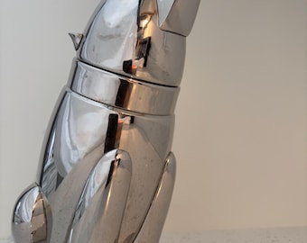 May include: A polished silver-toned metal cocktail shaker shaped like a sitting dog. The shaker features a smooth, reflective surface with a detachable head piece that serves as the lid, capturing the form of a canine looking upward.
