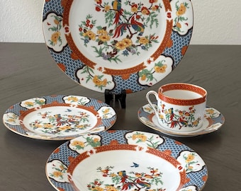 May include: A porcelain dinnerware set featuring a large plate, a smaller plate, a bowl, and a teacup with a saucer. Each piece displays a colorful bird and floral motif with a blue scale pattern border, orange geometric accents, and gold trim.