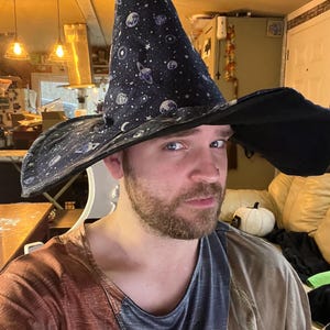 May include: A black wizard's hat with a wide brim and a tall, pointed crown. The hat features a pattern of white stars and planets. The person in the image is wearing a brown and blue shirt.