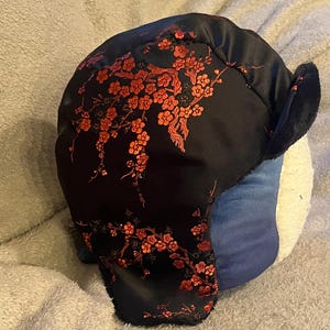 May include: Black winter hat with a red floral pattern. The hat has a faux fur trim around the ear flaps.