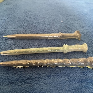 May include: Three resin pens with wand-like designs. The pens are in shades of brown, green, and gray. Each pen has a unique textured pattern and a pointed tip for writing. The pens are displayed on a blue surface.