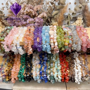 May include: A collection of gemstone chip bracelets in various colors, including pink, purple, blue, green, and red. The bracelets are displayed on wooden shelves, with dried flowers in the background. The brand name "ClassicBodhiCrystal" is visible.