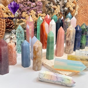 May include: A selection of multi-coloured crystal towers in various sizes and shades, including pink, green, blue, and clear. Some crystals are displayed on a small, square, beige platform. The background features dried flowers and a logo that reads "Glass RadhiCrystal".