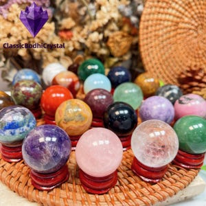 Può includere: Una collezione di sfere di cristallo colorate su piccoli supporti di legno rossi, esposte su un vassoio intrecciato. Le sfere variano di colore, tra cui rosa, viola, verde e blu. Il testo "ClassicBodhiCrystal" è visibile.