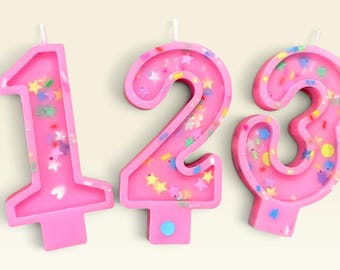 May include: Three pink wax candles shaped as the numbers one, two, and three. Each candle features a raised border and is filled with a variety of colorful, small confetti-like shapes including stars, circles, and hearts embedded in the wax.