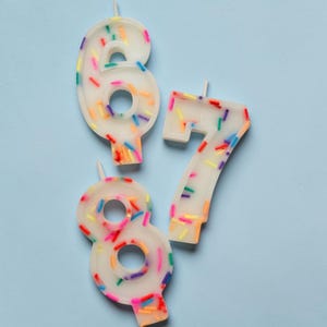 May include: Three white wax birthday candles shaped as the numbers six, seven, and eight. Each candle features a translucent finish embedded with small, colourful sprinkles in shades of red, blue, green, yellow, and purple, with a small wick at the top.