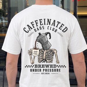 Op de afbeelding: Wit T-shirt met een afbeelding op de achterkant met de tekst "Caffeinated Dads Club" boven een skelet dat koffie uit een espressopotje giet. De tekst "Brewed Under Pressure" en "Est. 2020" staan ook op het shirt.